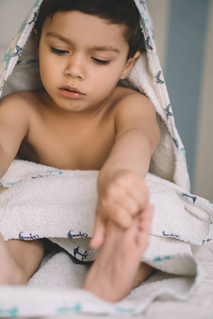 foyer sélectif de l'enfant mignon, recouvert d'une serviette à capuchon, toucher les pieds tout en étant assis sur le lit
 - Photo, image