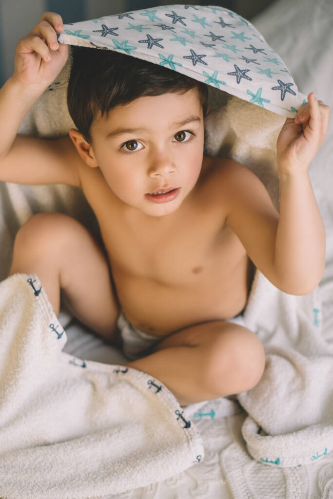 vue aérienne du garçon mignon, enveloppé dans une serviette à capuche, assis sur le lit et regardant la caméra
 - Photo, image