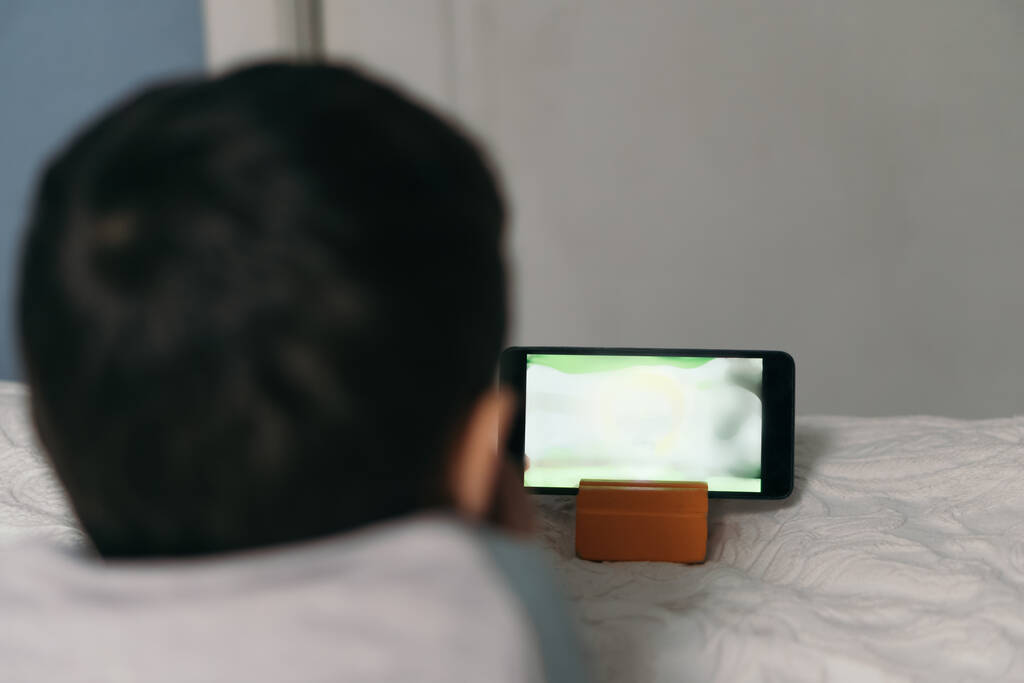 вид сзади ребенка, смотрящего обучающий мультфильм на смартфоне, лежа на кровати
 - Фото, изображение