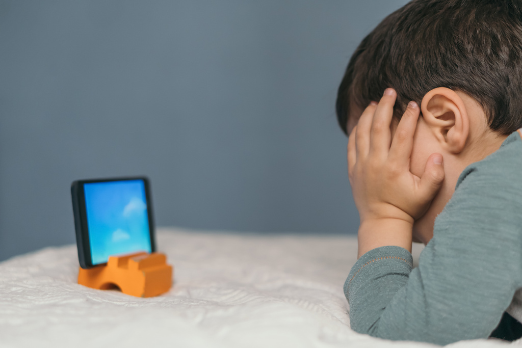 Kleiner Junge berührt Kopf, während er auf dem Smartphone Zeichentrickfilm anschaut, während er auf dem Bett liegt - Foto, Bild