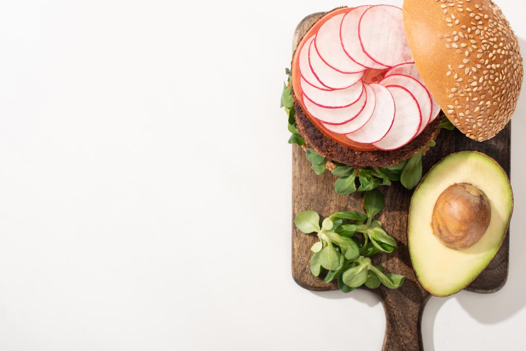 bovenaanzicht van heerlijke veganistische hamburger met radijs, avocado en greens op houten snijplank op witte achtergrond - Foto, afbeelding