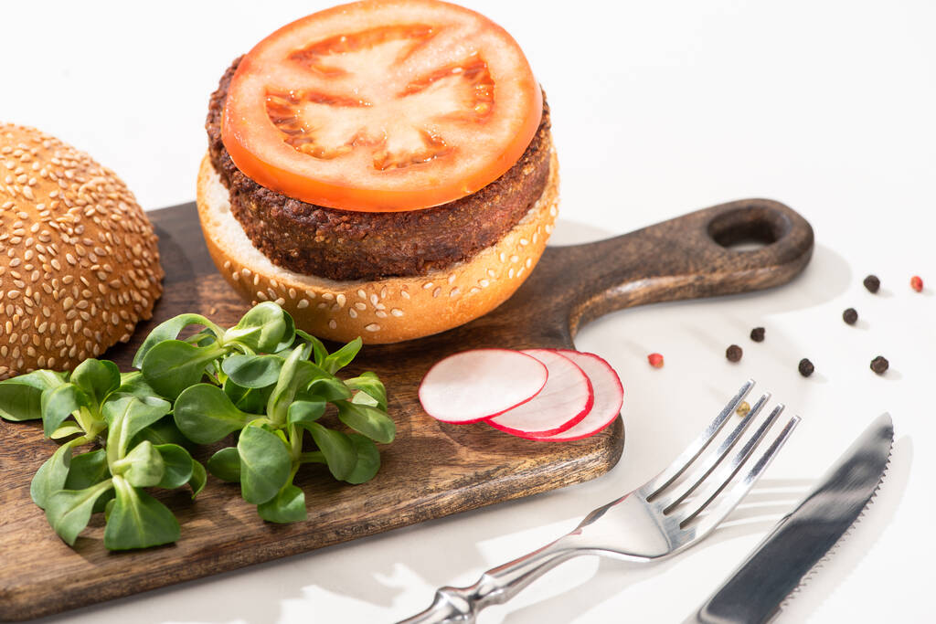 selektiver Schwerpunkt leckerer veganer Burger mit Rettich, Tomate und Mikrogemüse auf Holzschwein mit schwarzem Pfeffer in der Nähe von Gabel und Messer auf weißem Hintergrund - Foto, Bild