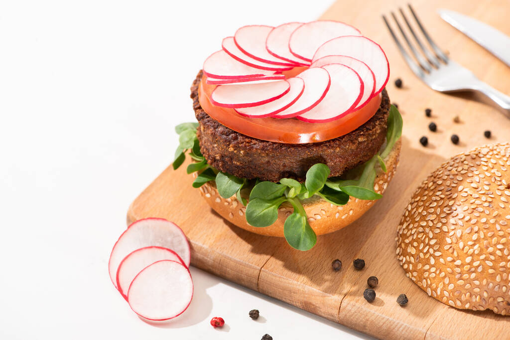 selective focus of delicious vegan burger with radish, tomato and microgreens on wooden boar with black pepper near fork and knife on white background - Photo, Image