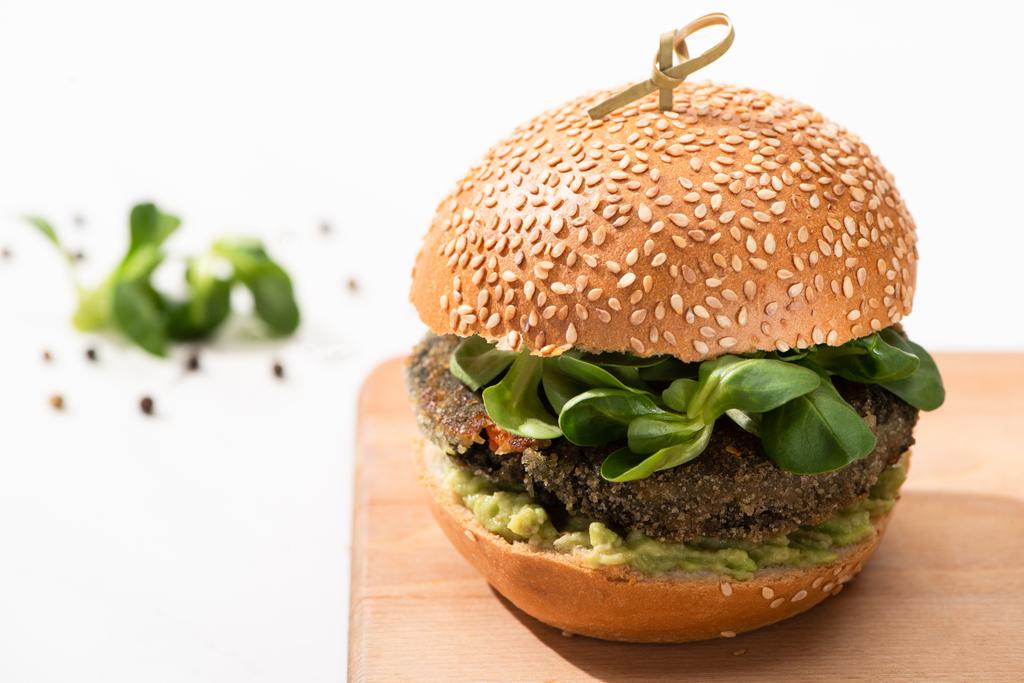 селективный фокус вкусного зеленого веганского бургера с микрозеленью и пюре авокадо на деревянном кабане с черным перцем на белом фоне
 - Фото, изображение