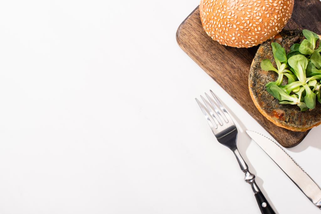 bovenaanzicht van veganistische hamburger met microgreens op houten bord bij bestek op witte achtergrond - Foto, afbeelding