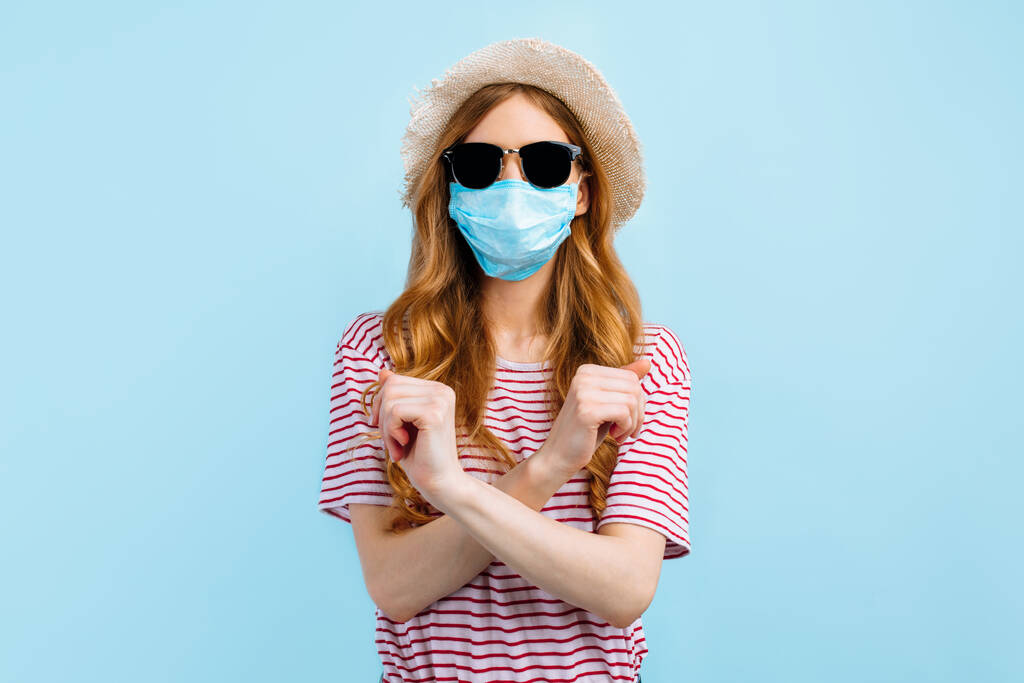 Egy fiatal lány nyári kalapban és napszemüvegben orvosi védőmaszkot visel, és egy megállító mozdulatot mutat keresztbe tett karokkal a kék háttéren. A karantén, a koronavírus fogalma - Fotó, kép