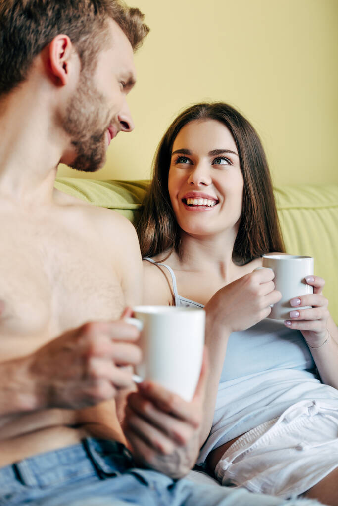 selective focus of cheerful couple holding cups with coffee and looking at each other in bedroom  - Photo, Image
