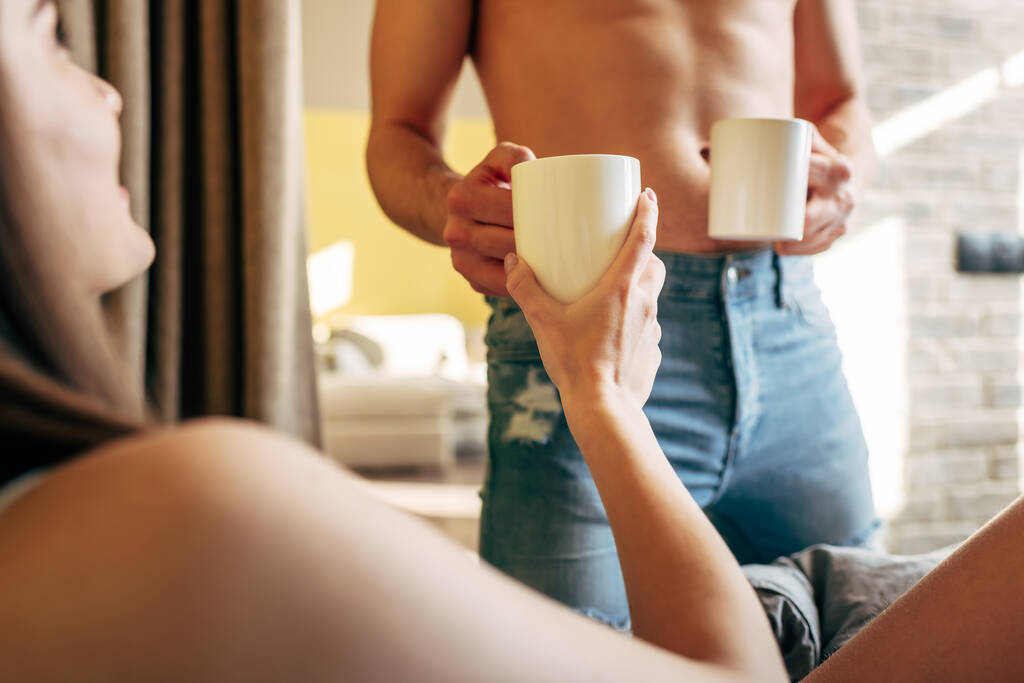 enfoque selectivo de hombre muscular sosteniendo tazas cerca de mujer atractiva en el dormitorio
  - Foto, imagen
