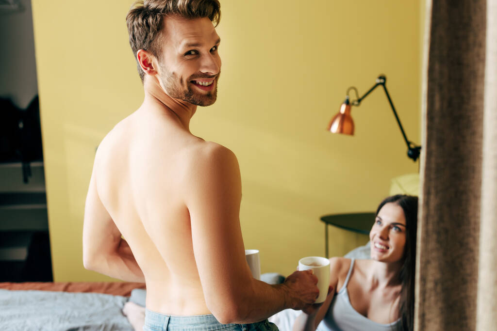 selective focus of shirtless man smiling while holding cups near girl in bedroom  - Photo, Image