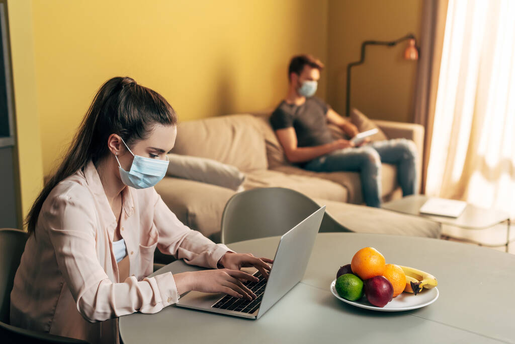 επιλεκτική εστίαση του ελεύθερου επαγγελματία στην ιατρική μάσκα χρησιμοποιώντας φορητό υπολογιστή κοντά στον άνθρωπο με ψηφιακή ταμπλέτα στο σπίτι  - Φωτογραφία, εικόνα