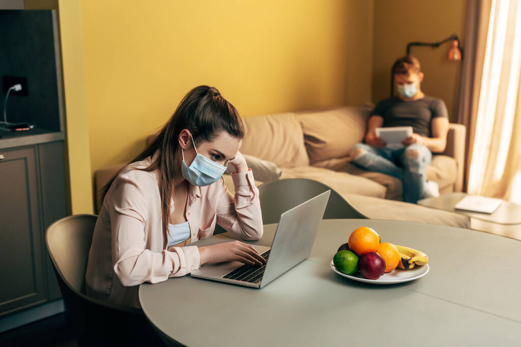 selektywne skupienie freelancera w masce medycznej przy użyciu laptopa w pobliżu owoców i człowieka z tabletu cyfrowego w domu  - Zdjęcie, obraz