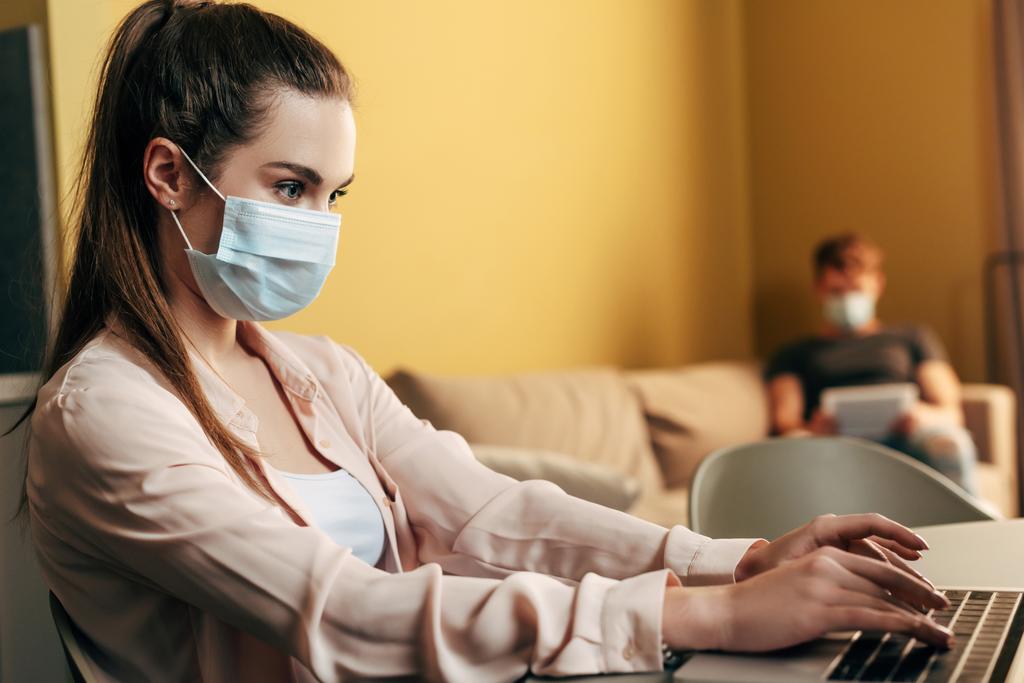 selektywny ostrość freelancer w masce medycznej wpisując na laptopie w pobliżu człowieka w domu  - Zdjęcie, obraz
