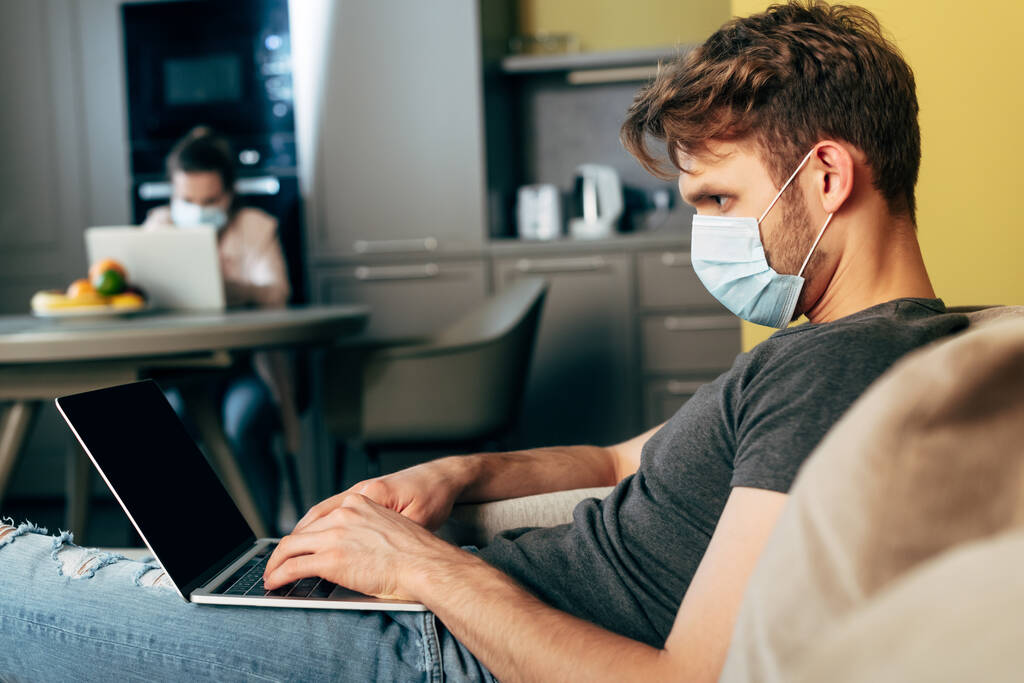 selective focus of freelancer in medical mask using laptop with blank screen near girlfriend at home - Photo, Image