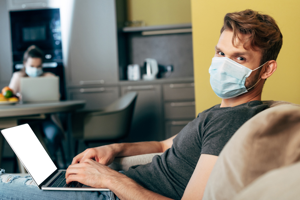 επιλεκτική εστίαση του ελεύθερου επαγγελματία στην ιατρική μάσκα χρησιμοποιώντας φορητό υπολογιστή με λευκή οθόνη κοντά φίλη  - Φωτογραφία, εικόνα