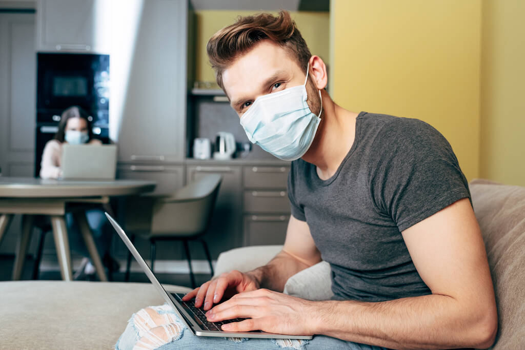 selective focus of freelancer in medical mask near laptop and girlfriend at home - Photo, Image