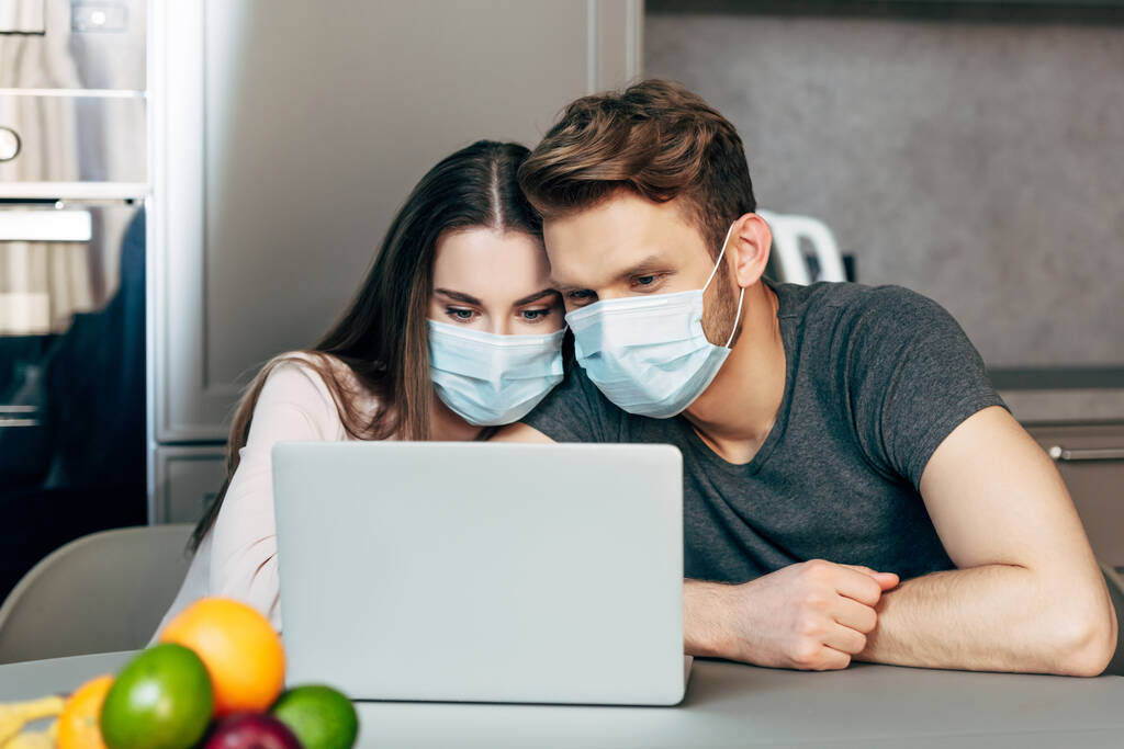 Selektiver Fokus des Paares in medizinischen Masken, das Film auf Laptop in der Nähe von Früchten anschaut  - Foto, Bild