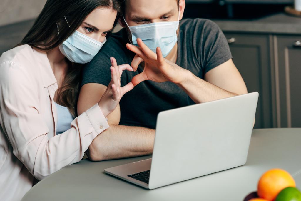 selective focus of couple in medical masks showing heart with hands while having video chat  - Photo, Image