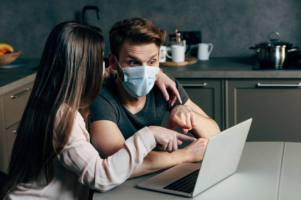 mujer señalando con el dedo a la computadora portátil cerca de novio en máscara médica
  - Foto, imagen