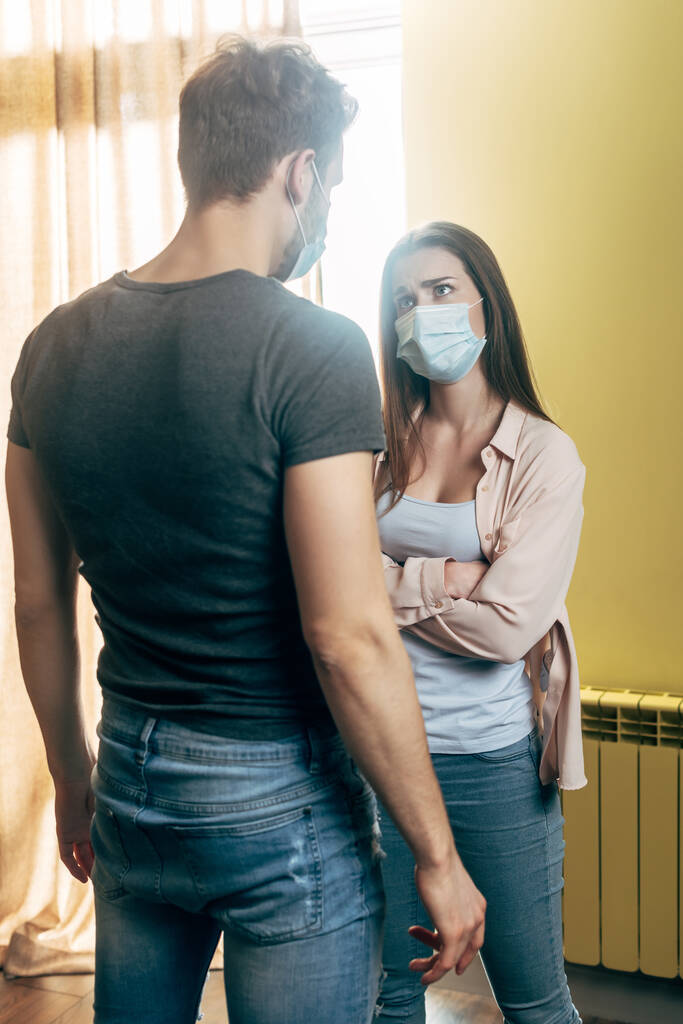 woman in medical mask standing with crossed arms while quarreling with boyfriend at home - Photo, Image