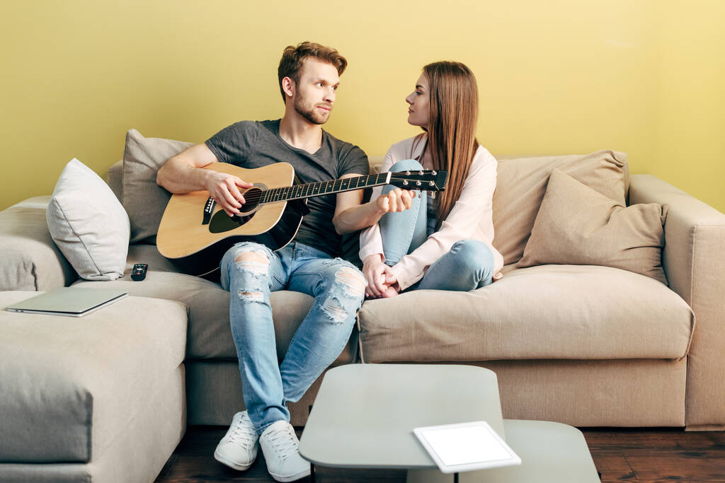 komea mies pelaa akustista kitaraa lähellä houkutteleva tyttö ja digitaalinen tabletti valkokankaalla
 - Valokuva, kuva
