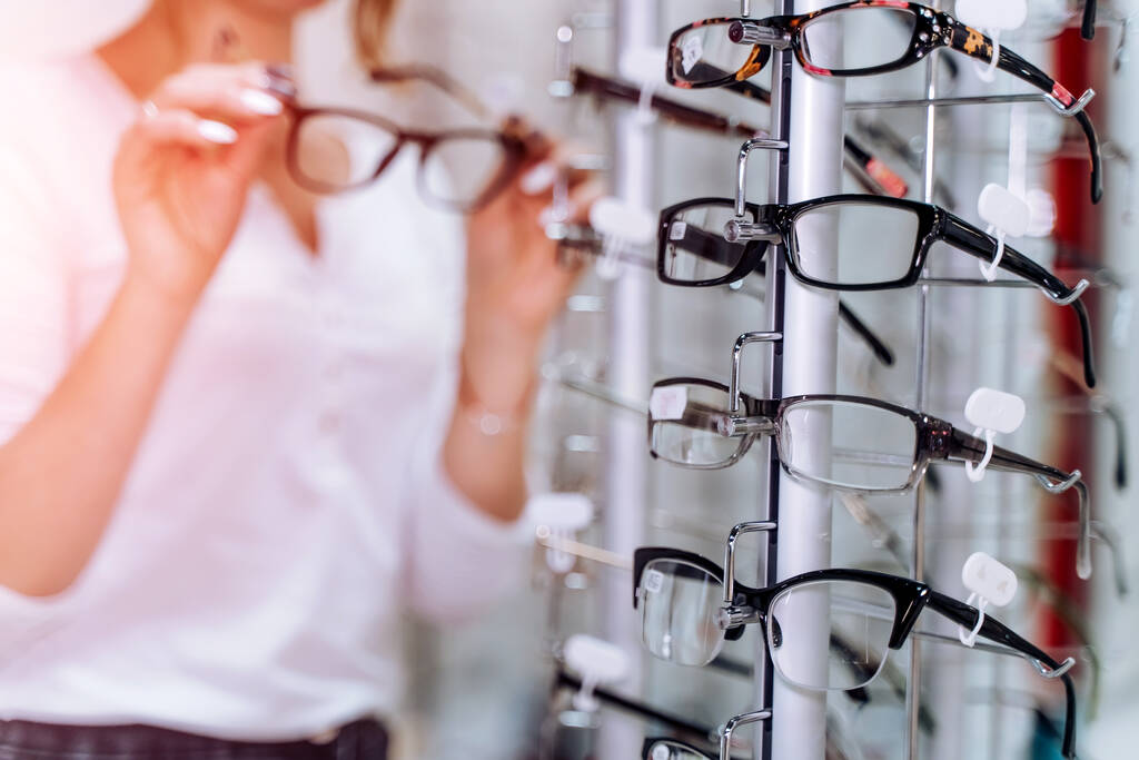 Female is standing with glasses in hands in the background Optical shop. Stand with spectacles. Eyesight correction. Close up. - Photo, Image
