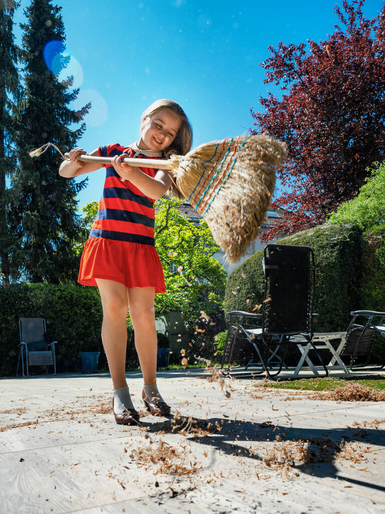 Μια μαθήτρια ποζάρει με μια σκούπα και με τα παπούτσια της μητέρας της στην αυλή. Σκούπισμα στον κήπο. Ηλιοφάνεια. Χαρούμενη παιδική ηλικία. - Φωτογραφία, εικόνα