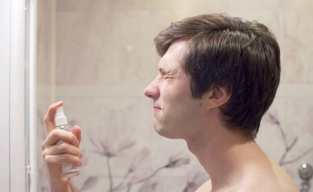 Κοντινό πορτρέτο του νεαρού όμορφου άντρα ψεκάζει λοσιόν ή αφτερσέιβ στο πρόσωπό του στο φωτεινό μπάνιο. Φροντίδα προσώπου μετά το ξύρισμα - Φωτογραφία, εικόνα