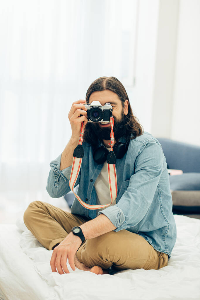 Hombre con ropa casual sentado en la cama y sonriendo mientras toma fotos con su cámara de cine
 - Foto, Imagen