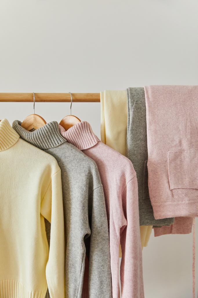vista de cerca de suéteres suaves de punto rosa, beige y gris y pantalones colgados en estante de madera aislados en blanco
 - Foto, Imagen