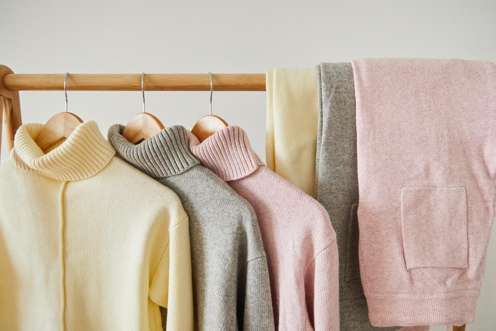 vista de cerca de suéteres suaves de punto rosa, beige y gris y pantalones colgados en estante de madera aislados en blanco
 - Foto, imagen