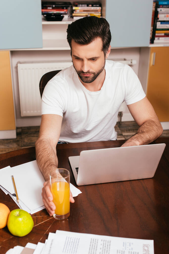 Selektivní zaměření pohledný muž drží sklenici pomerančového džusu při práci na notebooku doma  - Fotografie, Obrázek
