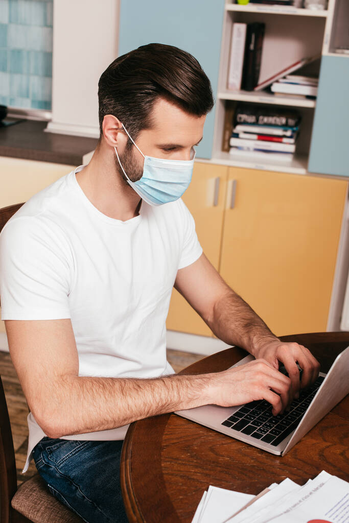 Άντρας με ιατρική μάσκα που χρησιμοποιεί φορητό υπολογιστή κοντά σε χαρτιά στο τραπέζι στο σπίτι  - Φωτογραφία, εικόνα
