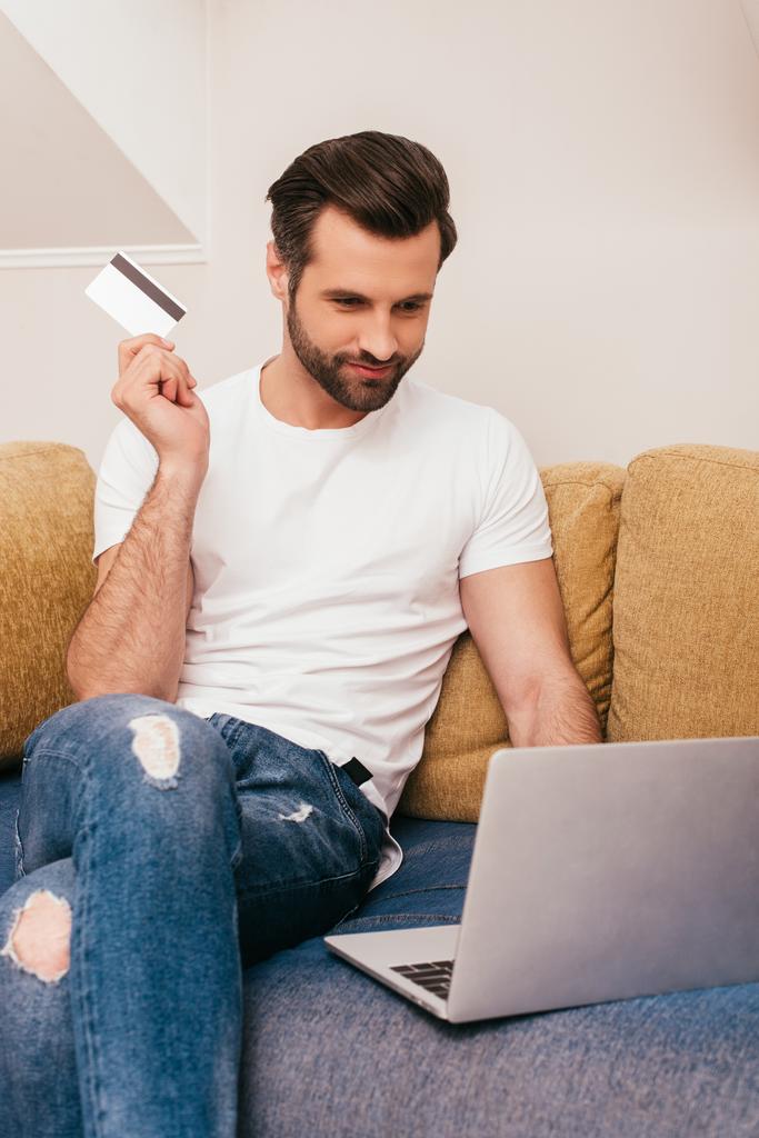 Beau homme tenant une carte de crédit et utilisant un ordinateur portable sur le canapé
  - Photo, image