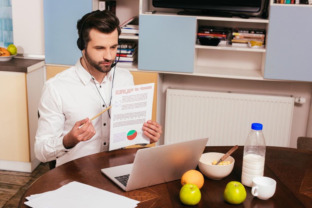 Freelancer in headset pointing on paper with charts while having video chat on laptop at home - Photo, Image