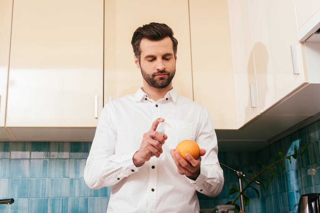 Низький кут зору красивого чоловіка, який тримає помаранчевий і ручний дезінфікуючий засіб на кухні
  - Фото, зображення