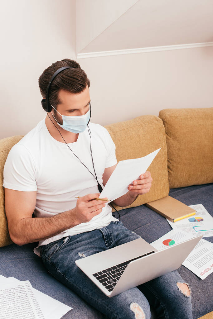 Freelancer en máscara médica y auriculares trabajando con papeles en sofá
  - Foto, Imagen