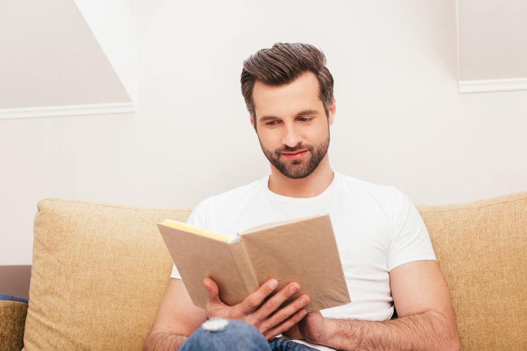 Selective focus of handsome man reading book on couch in living room - Photo, Image