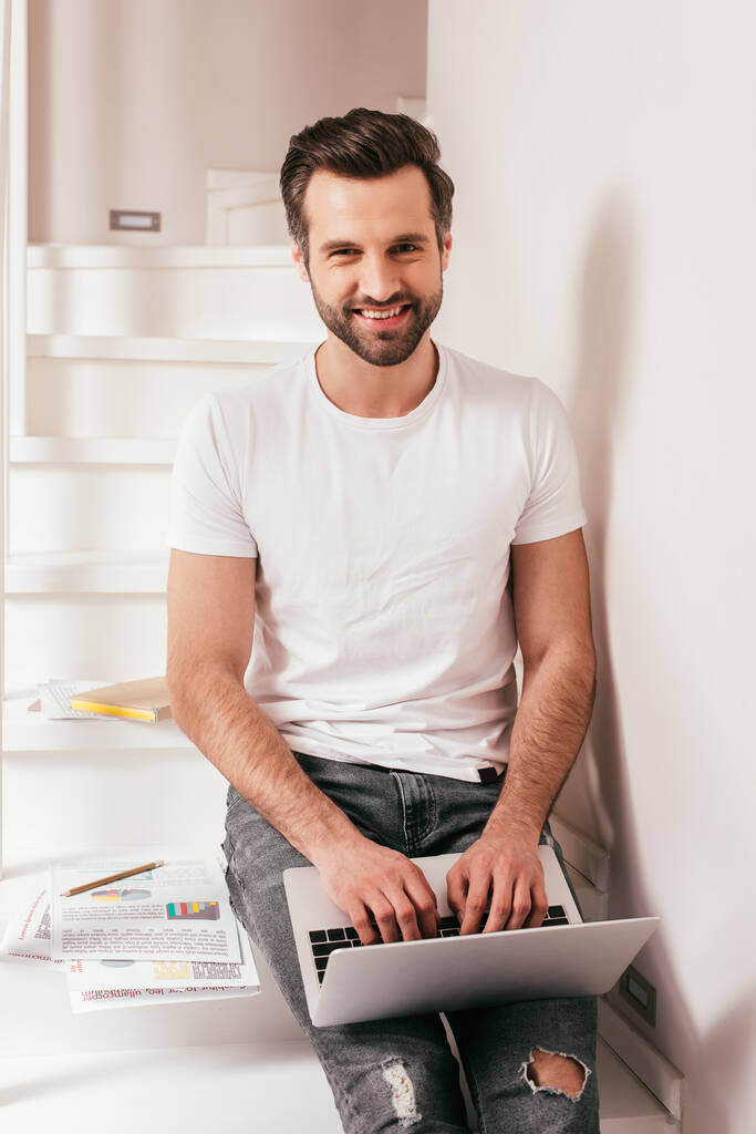 Bonito teletrabalhador sorrindo para a câmera ao usar laptop perto de documentos com gráficos em escadas
  - Foto, Imagem