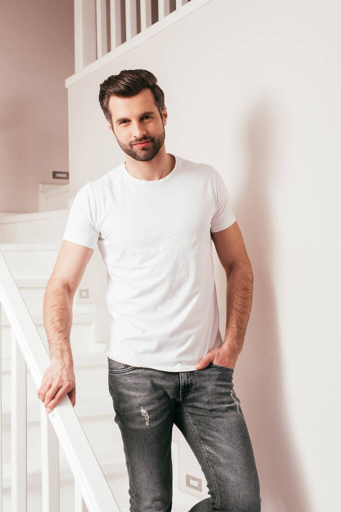 Красивий чоловік з рукою в кишені джинсів дивиться на камеру на сходах вдома
  - Фото, зображення