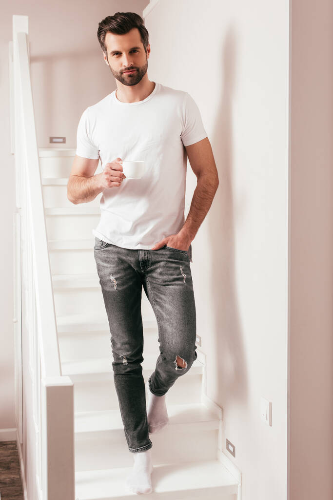 Jóképű férfi kezében egy csésze kávé és nézi a kamerát a lépcsőn otthon  - Fotó, kép