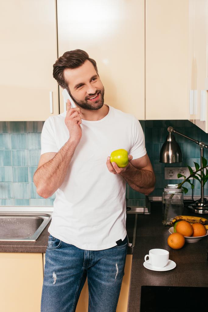 Uomo sorridente che parla su smartphone e tiene la mela in cucina
  - Foto, immagini