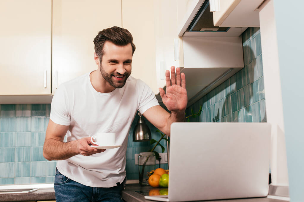 Выборочный фокус улыбающегося мужчины, держащего чашку кофе и имеющего видеозвонок на ноутбуке на кухне
  - Фото, изображение