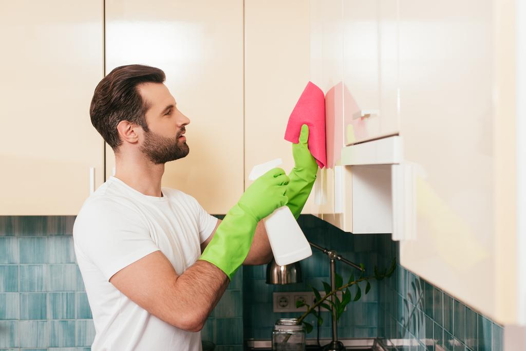 Oldalnézet jóképű férfi használ mosószer és rongy, miközben takarítás szekrény a konyhában  - Fotó, kép