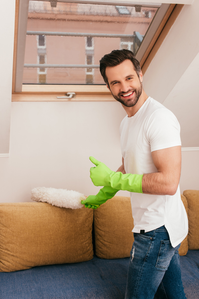 Vista laterale dell'uomo in guanti di gomma che mostra come gesto durante la pulizia del divano con spazzola antipolvere a casa
  - Foto, immagini