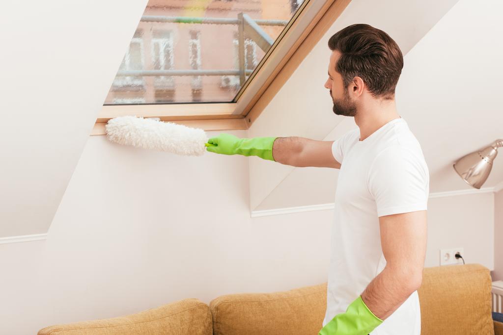 Vue latérale de l'homme en gants de caoutchouc nettoyage fenêtre avec brosse à poussière dans le salon
 - Photo, image