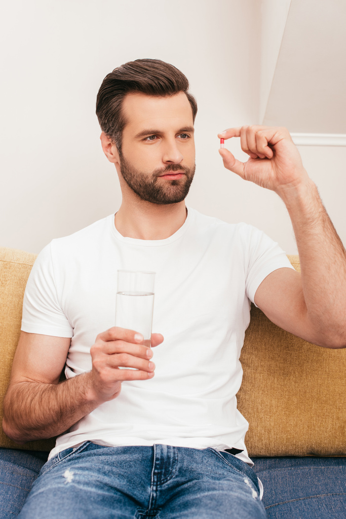 Komea mies pitelee lasillista vettä ja katselee pilleriä sohvalla
  - Valokuva, kuva