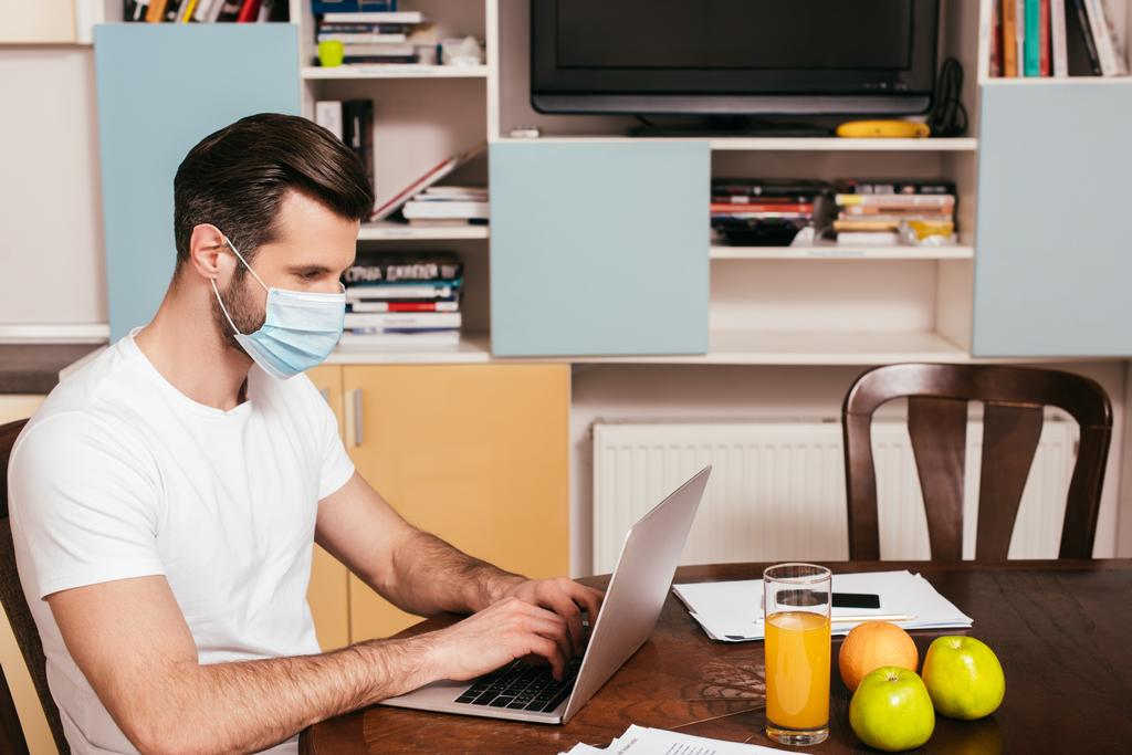 Widok z boku człowieka w masce medycznej pracy na laptopie w pobliżu soku pomarańczowego i owoców na stole  - Zdjęcie, obraz
