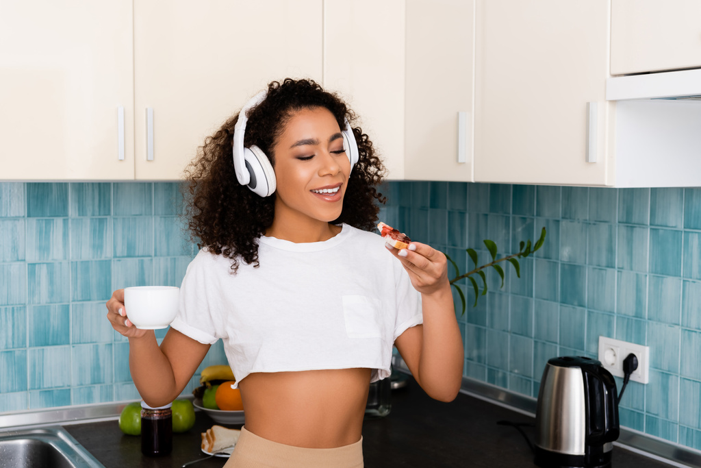 Χαρούμενη Αφροαμερικανή με ασύρματα ακουστικά που κρατάει τοστ με μαρμελάδα και καφέ.  - Φωτογραφία, εικόνα