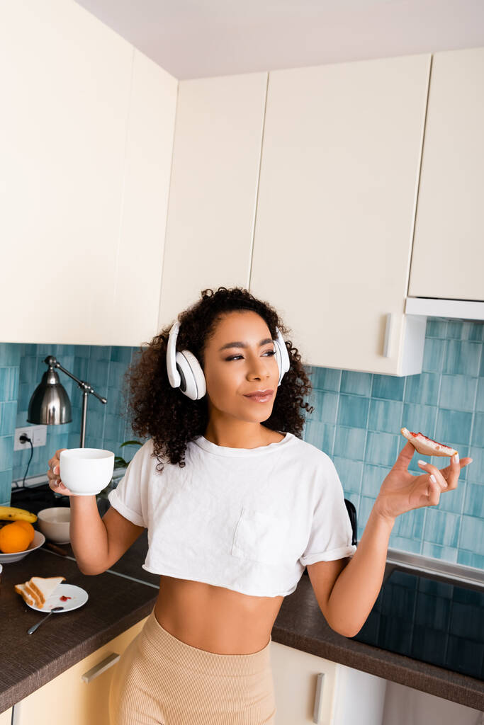 frisée afro-américaine fille dans des écouteurs sans fil tenant pain grillé avec confiture et tasse de café
  - Photo, image