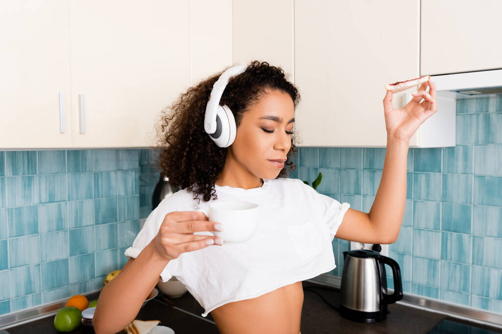 attraente ragazza afro-americana in cuffie wireless con pane tostato con marmellata e tazza di caffè
  - Foto, immagini